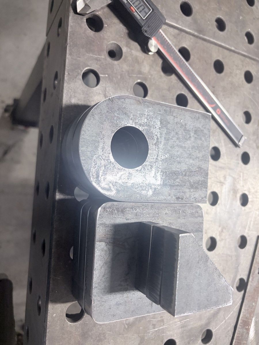 Thick steel plate lug and gusset parts on a fixture table next to a digital caliper.